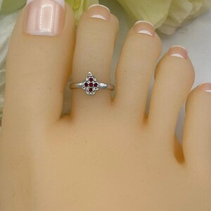 May include: A silver-toned toe ring featuring a four-leaf clover design with small, round, red gemstones. The ring is worn on a foot with a light skin tone, with a white background and floral accents.