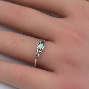 May include: A silver ring with an oval, light blue gemstone. The ring has a delicate, swirling design on the band. The ring is on a person's finger, with white flowers in the background.