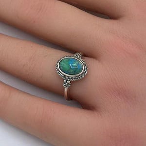 May include: A silver ring with an oval, blue-green stone. The ring has a detailed, beaded border around the stone and a thin band. The ring is displayed on a hand with manicured nails, with white and yellow flowers in the background.