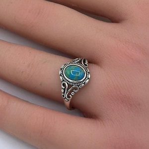 May include: A silver ring with an oval, turquoise-colored stone. The ring has an ornate, filigree design around the stone. The ring is on a hand with manicured nails, with white and pink polish. The background includes white and yellow flowers.
