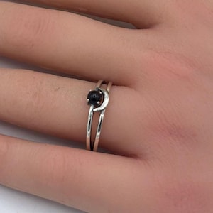 Op de afbeelding: Een zilveren ring met een kleine, ronde, zwarte steen gezet op een gesplitste band. De ring zit aan een hand met een French manicure. Witte bloemen zijn op de achtergrond.