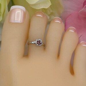 May include: A silver toe ring with a heart-shaped, red gemstone is worn on a foot. The ring is delicate and sits on the second toe. The background features white, yellow, and pink flowers.