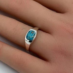 Puede incluir: Un anillo de plata con una piedra turquesa ovalada. El anillo tiene un acabado pulido y un diseño sencillo y elegante. La piedra turquesa tiene un patrón único y está engastada en un bisel.