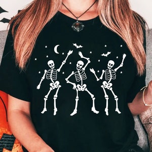 Könnte beinhalten: Schwarzes T-Shirt mit einer weißen Grafik von drei tanzenden Skeletten. Die Skelette sind in einer festlichen, partyartigen Pose. Das Shirt hat ein sternenartiges Muster und einen Halbmond im Hintergrund.