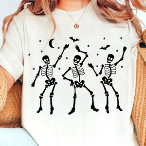 Könnte beinhalten: Weißes T-Shirt mit einem Motiv von drei Skeletten, die unter einem Halbmond und Sternen tanzen. Die Skelette sind schwarz und haben ein einfaches Design.
