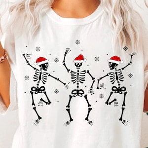 May include: White t-shirt with a graphic of three skeletons wearing Santa hats and dancing. The skeletons are black and white, and the hats are red and white.