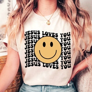 May include: A white t-shirt with a black graphic print of a yellow smiley face and the text "JESUS LOVES YOU" repeated multiple times.