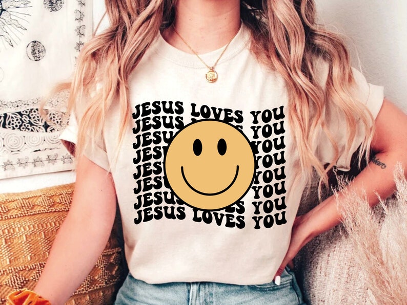 May include: A white t-shirt with a black graphic print of a yellow smiley face and the text "JESUS LOVES YOU" repeated multiple times.