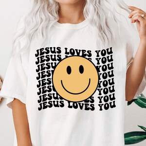 May include: White t-shirt with a black and yellow smiley face and the text "JESUS LOVES YOU" repeated in a retro font.