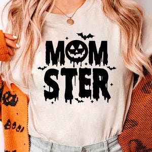 Op de afbeelding: Een wit T-shirt met een zwarte grafische afbeelding waarop "MOMSTER" staat, met een druipend pompoengezicht in het midden van het woord "MOM".