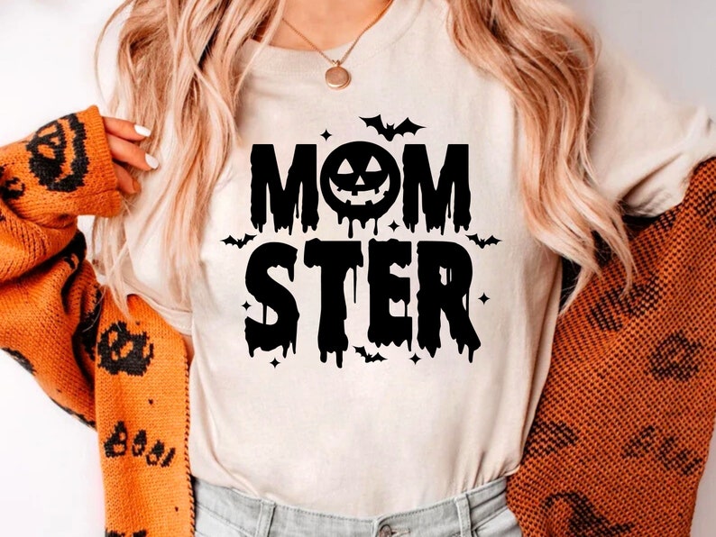 Op de afbeelding: Een wit T-shirt met een zwarte grafische afbeelding waarop "MOMSTER" staat, met een druipend pompoengezicht in het midden van het woord "MOM".