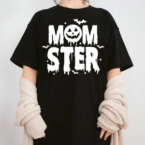 Op de afbeelding: Zwart T-shirt met witte tekst die "MOMSTER" luidt met een pompoengezicht in de "O". De tekst heeft een druppelend effect en er zijn kleine vleermuizen en sterren rond de tekst.