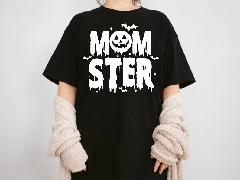 Op de afbeelding: Zwart T-shirt met witte tekst die "MOMSTER" luidt met een pompoengezicht in de "O". De tekst heeft een druppelend effect en er zijn kleine vleermuizen en sterren rond de tekst.