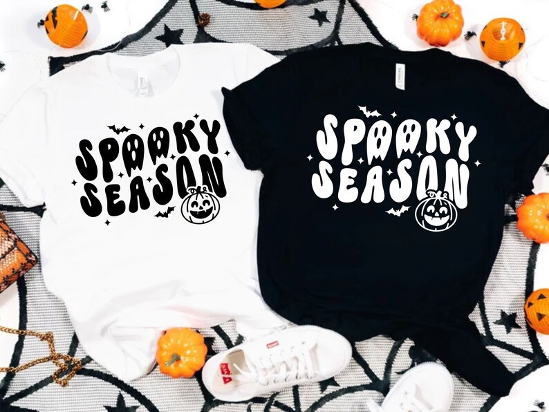 May include: Two t-shirts with the text "Spooky Season" in a retro font. The shirts are black and white and feature a pumpkin and bats.