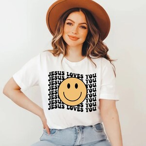 May include: White t-shirt with a yellow smiley face and the text "JESUS LOVES YOU" repeated in black.
