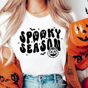 May include: A white t-shirt with the text "Spooky Season" in black, stylized lettering. The text is surrounded by small stars and bats. A cartoon pumpkin with a smiling face is below the text.