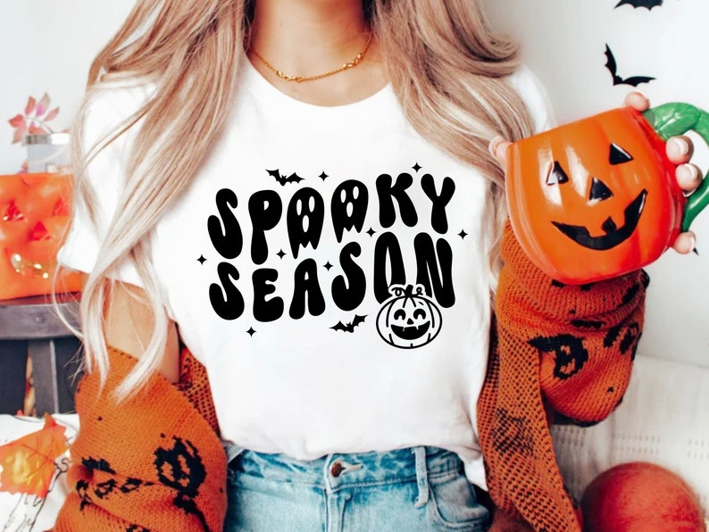 May include: A white t-shirt with the text "Spooky Season" in black, stylized lettering. The text is surrounded by small stars and bats. A cartoon pumpkin with a smiling face is below the text.