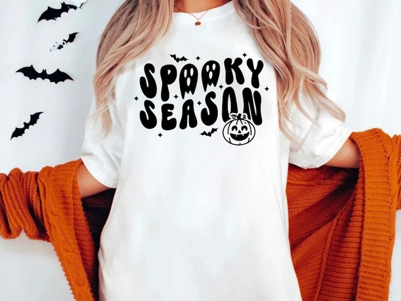 May include: A white t-shirt with a black graphic that says "Spooky Season" in a retro font. The graphic also includes a black silhouette of a pumpkin with a smiling face and bats.