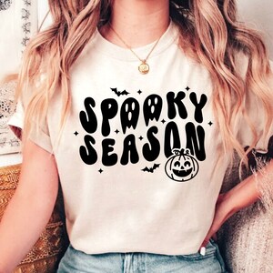 May include: A white t-shirt with a black graphic that says "Spooky Season" in a retro font. The graphic also includes a pumpkin with a smiling face and bats.