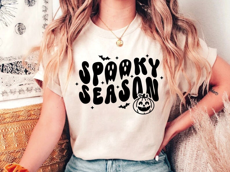 May include: A white t-shirt with a black graphic that says "Spooky Season" in a retro font. The graphic also includes a pumpkin with a smiling face and bats.