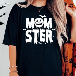 Op de afbeelding: Zwart T-shirt met een witte afbeelding die "MOMSTER" zegt met een druppelend effect. De afbeelding bevat een pompoengezicht en vleermuizen.