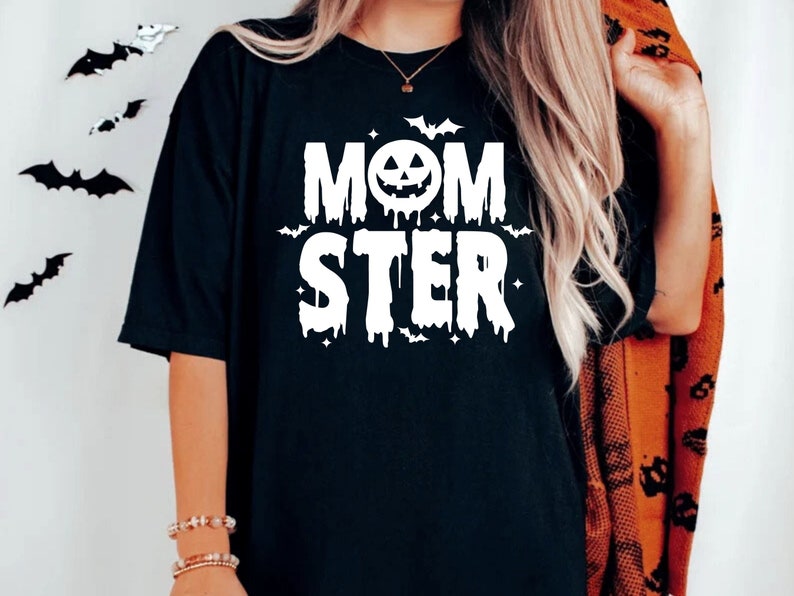 Op de afbeelding: Zwart T-shirt met een witte afbeelding die "MOMSTER" zegt met een druppelend effect. De afbeelding bevat een pompoengezicht en vleermuizen.