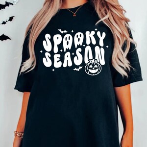 May include: Black t-shirt with white text that says "Spooky Season" in a retro font. The text is surrounded by small white stars and a white pumpkin with a smiling face.