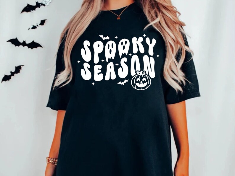 May include: Black t-shirt with white text that says "Spooky Season" in a retro font. The text is surrounded by small white stars and a white pumpkin with a smiling face.