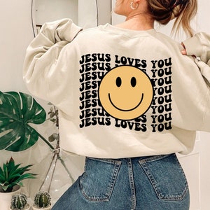 May include: A cream colored sweatshirt with a black and yellow smiley face graphic and the text "JESUS LOVES YOU" repeated multiple times.