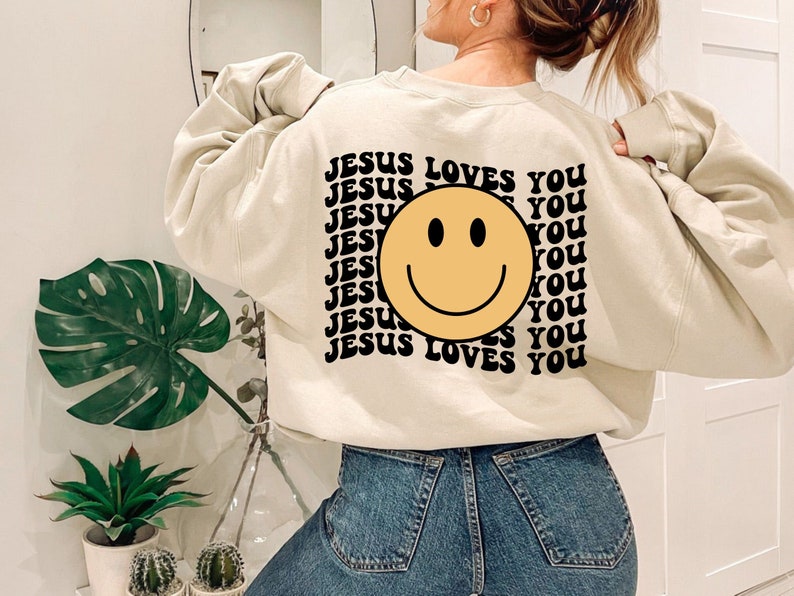 May include: A cream colored sweatshirt with a black and yellow smiley face graphic and the text "JESUS LOVES YOU" repeated multiple times.