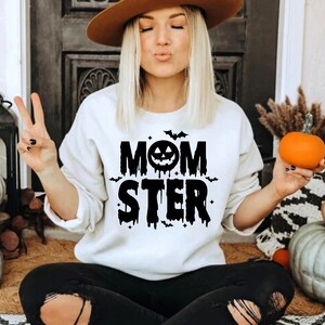 Op de afbeelding: Wit sweatshirt met zwarte tekst die "Momster" zegt met een zwart pompoengezicht en vleermuizen. Het sweatshirt wordt gedragen door een persoon met lang blond haar en een bruine hoed.