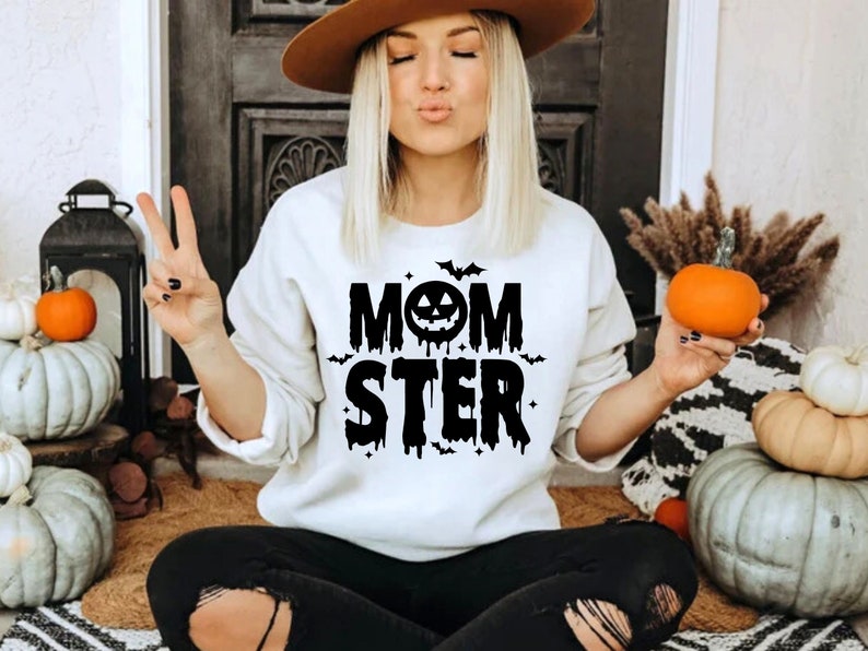Op de afbeelding: Wit sweatshirt met zwarte tekst die "Momster" zegt met een zwart pompoengezicht en vleermuizen. Het sweatshirt wordt gedragen door een persoon met lang blond haar en een bruine hoed.