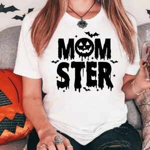 Op de afbeelding: Wit T-shirt met zwarte tekst die "Momster" luidt met een druppelend effect. De tekst bevat een zwarte pompoen met een jack-o-lantern gezicht.