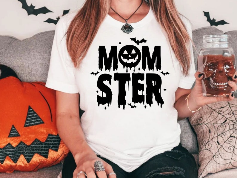 Op de afbeelding: Wit T-shirt met zwarte tekst die "Momster" luidt met een druppelend effect. De tekst bevat een zwarte pompoen met een jack-o-lantern gezicht.