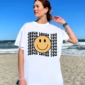 May include: White t-shirt with a black and yellow smiley face and the text "JESUS LOVES YOU" repeated in a retro pattern.