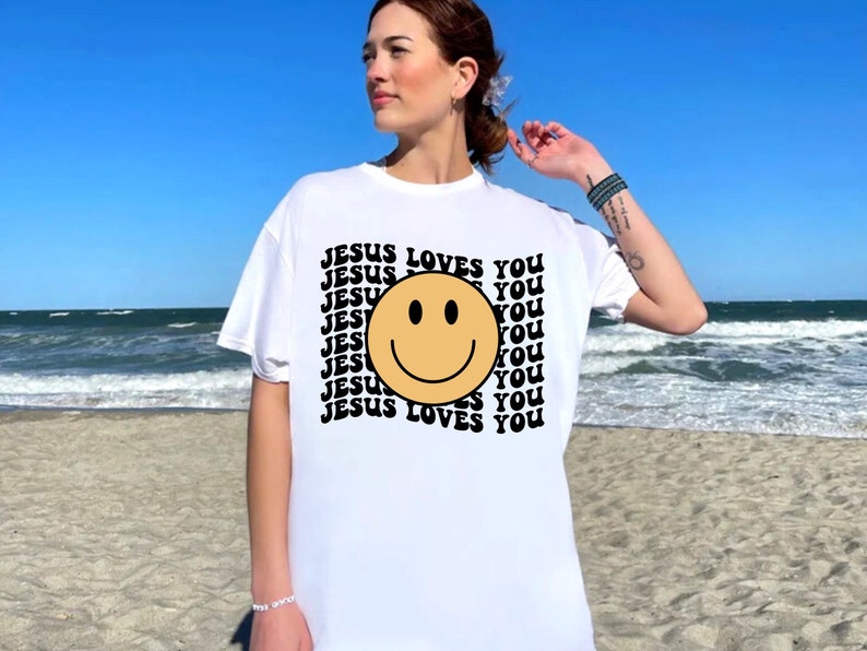 May include: White t-shirt with a black and yellow smiley face and the text "JESUS LOVES YOU" repeated in a retro pattern.