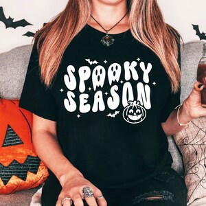May include: A black t-shirt with a white graphic that says "Spooky Season" in a retro font. The graphic also includes a cartoon ghost and a pumpkin.