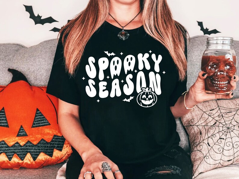 May include: A black t-shirt with a white graphic that says "Spooky Season" in a retro font. The graphic also includes a cartoon ghost and a pumpkin.