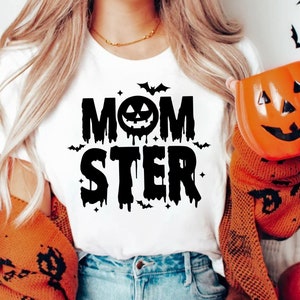 Op de afbeelding: Wit T-shirt met zwarte tekst die "MOMSTER" luidt met een druipend pompoengezicht in het midden van het woord "MOM".