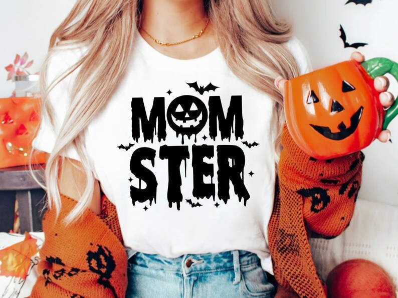 Op de afbeelding: Wit T-shirt met zwarte tekst die "MOMSTER" luidt met een druipend pompoengezicht in het midden van het woord "MOM".