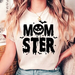 Op de afbeelding: Een wit T-shirt met een zwarte grafische afbeelding waarop "MOMSTER" staat met een druppelend effect en een pompoengezicht in de "O". Er zijn vleermuizen en sterren rond de tekst.