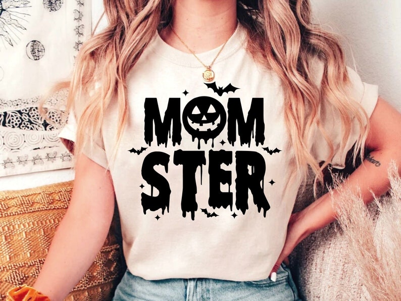 Op de afbeelding: Een wit T-shirt met een zwarte grafische afbeelding waarop "MOMSTER" staat met een druppelend effect en een pompoengezicht in de "O". Er zijn vleermuizen en sterren rond de tekst.