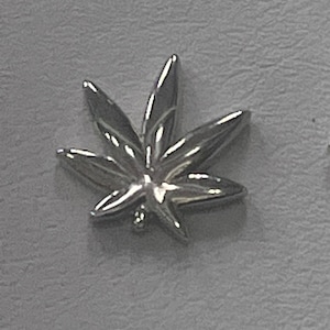 Puede incluir: Un dije plateado con forma de hoja de cannabis. El dije presenta un diseño detallado y realista con segmentos de hojas puntiagudos y un tallo central. La superficie refleja la luz, creando reflejos y sombras. Probablemente sea un colgante o un pendiente.
