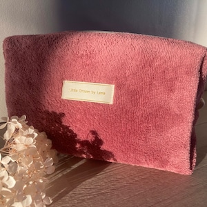 Può includere: Un beauty case rettangolare color rosa polveroso con una superficie morbida e strutturata. La borsa presenta una piccola etichetta rettangolare con la scritta "Little Dream by Lena" in lettere dorate. Un rametto di fiori bianchi secchi è in primo piano.