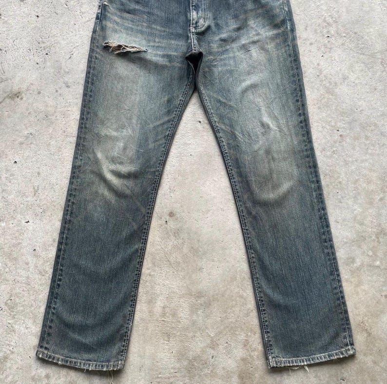 May include: Faded blue denim jeans with a distressed look. The jeans have a worn appearance with a tear on the upper left leg. The jeans have a classic five-pocket design and a straight leg cut.