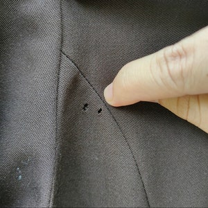 May include: Close-up of a dark brown fabric with two small holes near a seam. The fabric appears to be from a suit jacket or blazer, showing signs of wear or damage. A finger points towards the holes.
