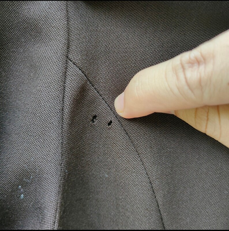 May include: Close-up of a dark brown fabric with two small holes near a seam. The fabric appears to be from a suit jacket or blazer, showing signs of wear or damage. A finger points towards the holes.