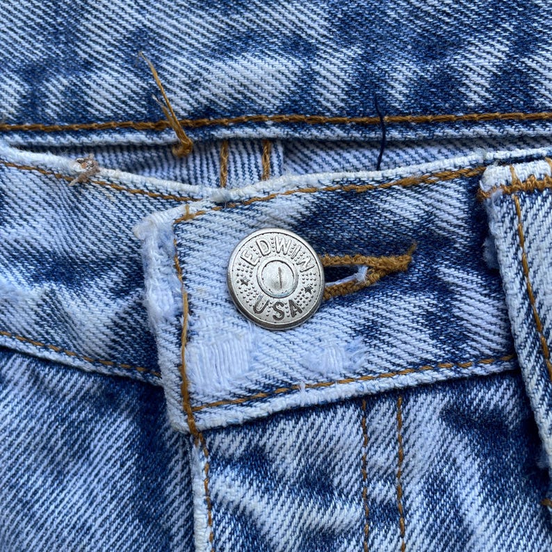 May include: Close-up of a light blue denim jeans button with the text "EDWIN USA" embossed on the silver button. The jeans have a faded, textured appearance with visible stitching.