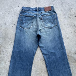 Pu&ograve; includere: Vista posteriore di jeans blu in denim con un classico design a cinque tasche. I jeans presentano un lavaggio sbiadito, una toppa in pelle marrone e il logo Levi's sulle tasche posteriori. I jeans sono realizzati in denim resistente.