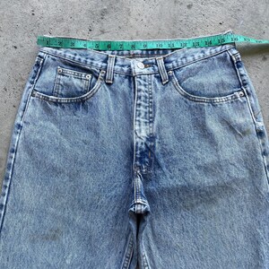 May include: Light blue denim jeans with a classic five-pocket design. The jeans have a button and zipper closure, belt loops, and a faded wash. A measuring tape is placed across the waist, showing a measurement of approximately 15 inches.